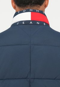 Chaqueta acolchada en color marino con collar alto y una banda de logo en rojo, blanco y azul. Textura suave con costuras en la parte posterior.