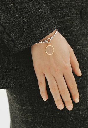 Mano que lleva una pulsera de cuentas plateadas con un encanto dorado redondo, descansando junto a la manga de un traje negro texturizado con botones.
