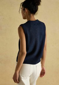 Tumesinist värvi varrukateta kampsun koos soonikkoelise krae ja äärega, tekstuurse kootud kangaga, kantud valgete pükste peal, lihtsa disainiga.