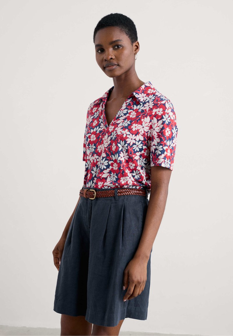 Red floral blouse with white and blue accents, collared neckline, and short sleeves paired with dark pleated shorts and a braided belt.