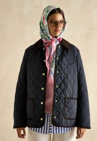 Granatowa pikowana kurtka z brązowym kołnierzem z sztruksu, złotymi zatrzaskami i brązowym wykończeniem, nałożona na różowy sweter i w paski koszulę.