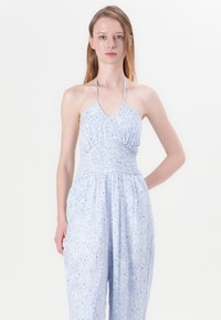 Hvit jumpsuit med blå blomsterdesign, halterneck, smocket bærestykke og vide ben. Lett stoff med en myk tekstur.