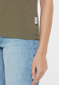 Olive green cotton t-shirt with a round neckline, short sleeves, side logo tag, paired with light blue denim jeans. Smooth texture.