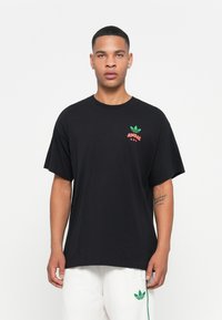 T-shirt en coton noir à manches courtes. Présente un logo Adidas vert et rouge sur la poitrine. Design simple, coupe décontractée et col rond.