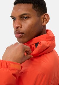 Giacca arancione brillante con colletto alto, polsi regolabili e tessuto testurizzato. Il modello tiene il colletto con una mano.