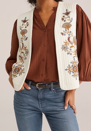 Vrouw draagt een wit gewatteerd vest met blauwbruine bloemrijke borduursels over een roestkleurige blouse met knopen en een blauwe spijkerbroek met een zwarte riem.