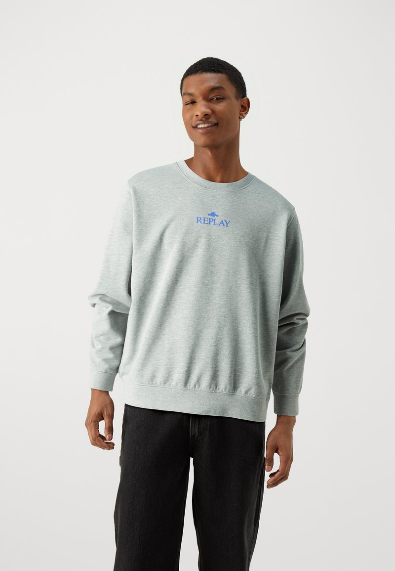 Joven sonriendo, vistiendo un sudadera gris claro con el logo "REPLAY" y pantalones negros, de pie frente a un fondo blanco liso.