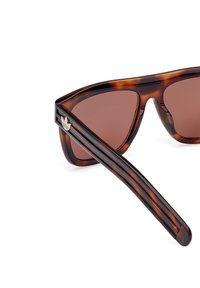 Ochelari de soare tortoiseshell maro cu un cadru triunghiular, lentile gradient maro și logo accent pe argintiu pe braț.