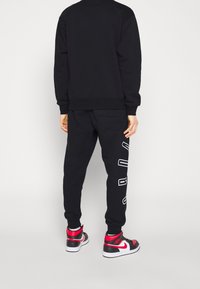 Sudadera negra y pantalones deportivos mostrados desde atrás, con puños elásticos y texto grande impreso en los pantalones, combinados con zapatillas rojas y negras.