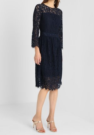 Femme portant une robe en dentelle bleu marine longueur genou avec des manches trois-quarts et des sandales à talons hauts couleur beige sur un fond uni.