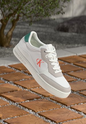 Zapatillas de cuero blanco con acentos de ante gris y un talón en color azul verde. Presentan un logo naranja brillante y una suela texturizada, posicionadas sobre baldosas de madera.