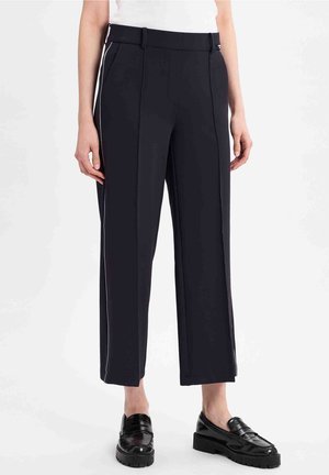 Kvinde iført sorte korte bukser med hvide sidelommer og sorte skinnende loafers, stående mod en ensfarvet hvid baggrund.