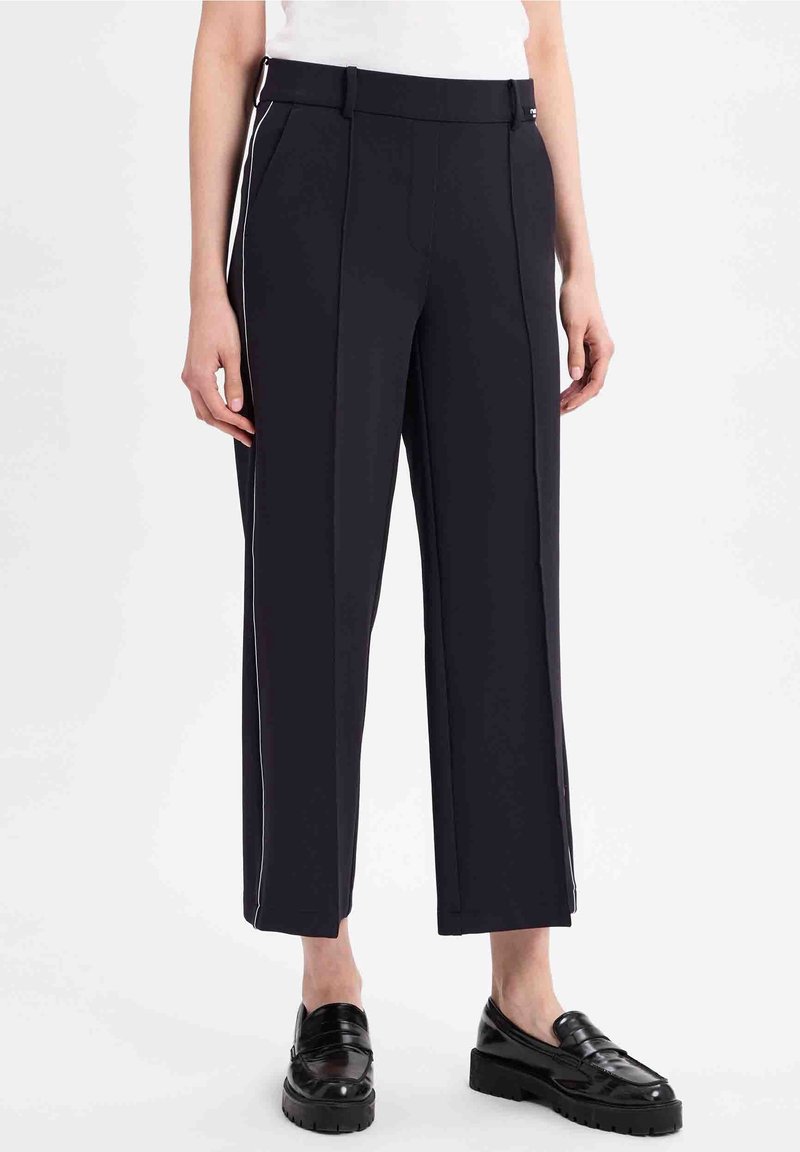 Femme portant un pantalon noir court avec des bandes blanches sur les côtés et des mocassins noirs brillants, debout devant un fond blanc uni.