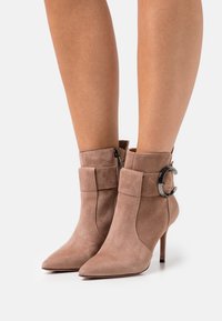 Bottines en daim beige avec un bout pointu, dotées d'un accent circulaire en argent et d'une fermeture éclair sur le côté, reposant sur un talon haut élancé.