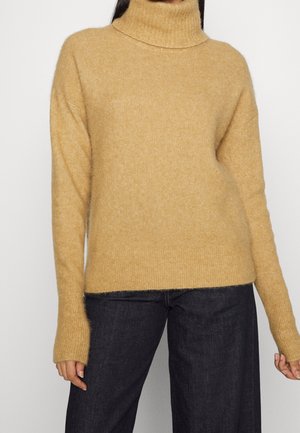 Femme portant un pull à col roulé couleur chameau à manches longues et un pantalon large foncé, debout devant un fond uni.
