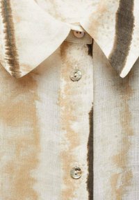 Primo piano di un colletto di una maglietta tie-dye beige e marrone con due bottoni iridescenti e dettaglio della texture del tessuto.