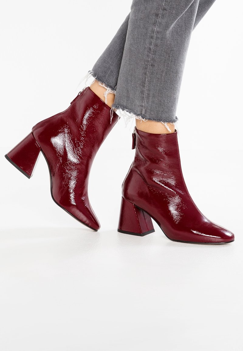 Bottines rouge foncé brillantes à talons carrés portées avec un jean gris à ourlets effilochés sur fond blanc.