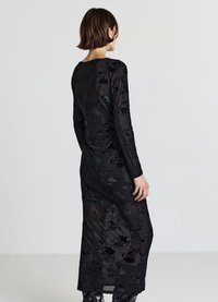 Robe longue noire avec un motif floral en velours, dotée de manches longues et d'une texture légèrement transparente. Le design est élégant et ajusté.