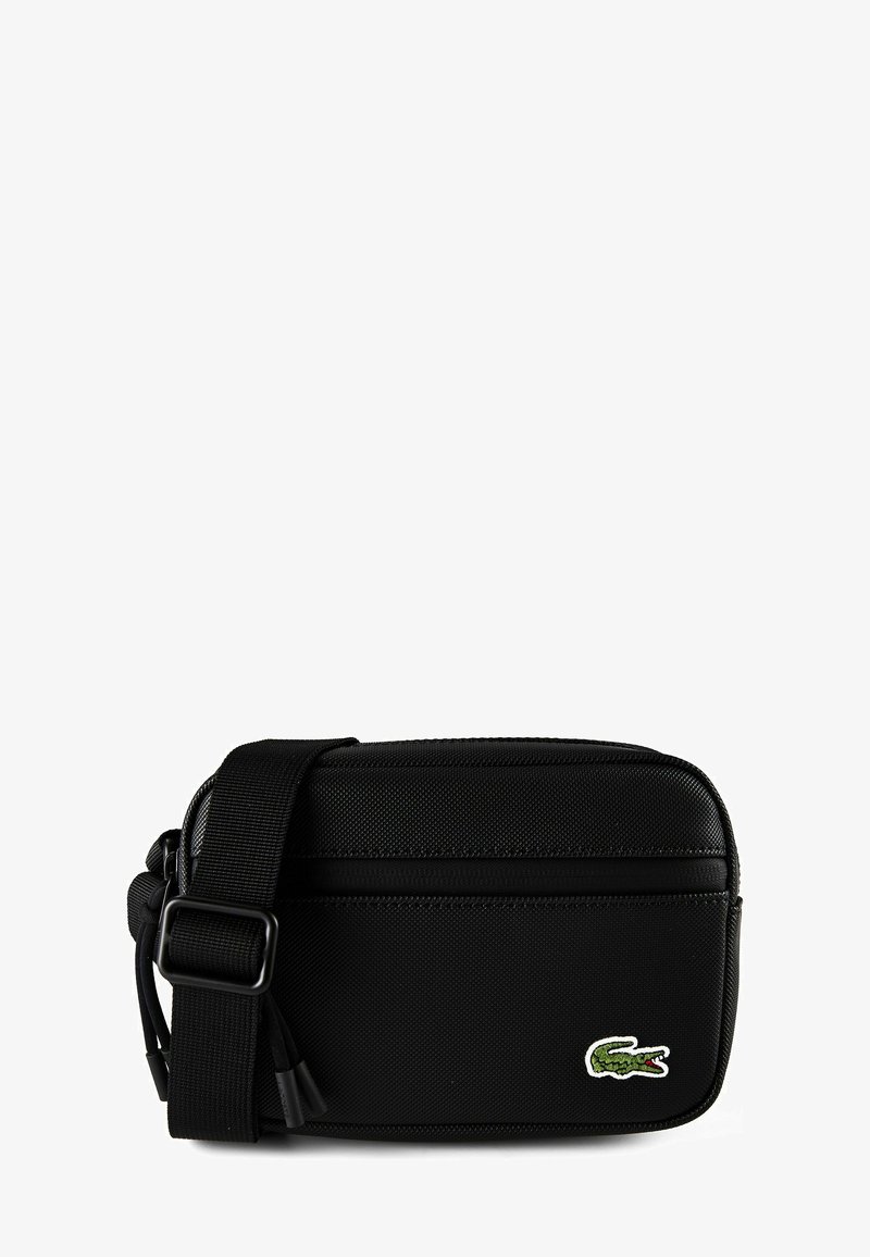 Bolso negro de bandolera con correa ajustable y bolsillo frontal con cremallera, que presenta un pequeño logo de cocodrilo verde en la esquina inferior derecha.