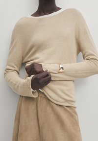 Pull en tricot beige à manches longues avec un large décolleté et une texture côtelée, porté par-dessus un haut blanc, associé à une jupe en daim couleur sable.