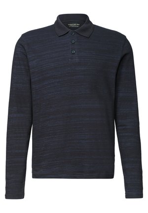 Mann mit dunklem lockigem Haar und Bart, der ein dunkles langärmeliges Polohemd mit weißem Unterhemd vor einem schlichten Hintergrund trägt.