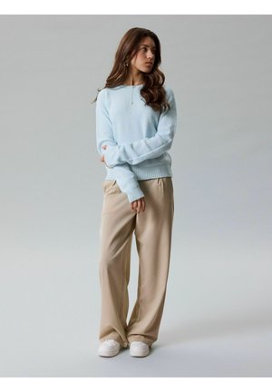 Jeune femme aux longs cheveux bruns portant un pull bleu clair, un pantalon beige à jambes larges et des baskets blanches, debout les bras croisés.