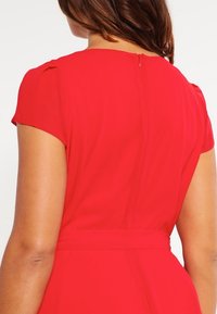 Robe rouge à manches courtes, taille cintrée et fermeture éclair dissimulée dans le dos. Tissu lisse et design épuré et minimaliste.