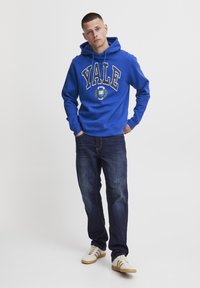 Felpa blu con la scritta "YALE" stampata in lettere bianche, caratterizzata da un logo circolare con dettagli di alloro. Abbinata a jeans scuri e sneaker chiare.