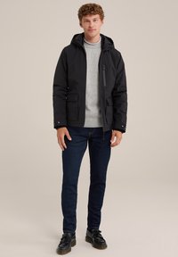 Chaqueta negra impermeable con capucha, bolsillo con cremallera y dos bolsillos frontales con solapa, llevada sobre un suéter gris claro y jeans oscuros.