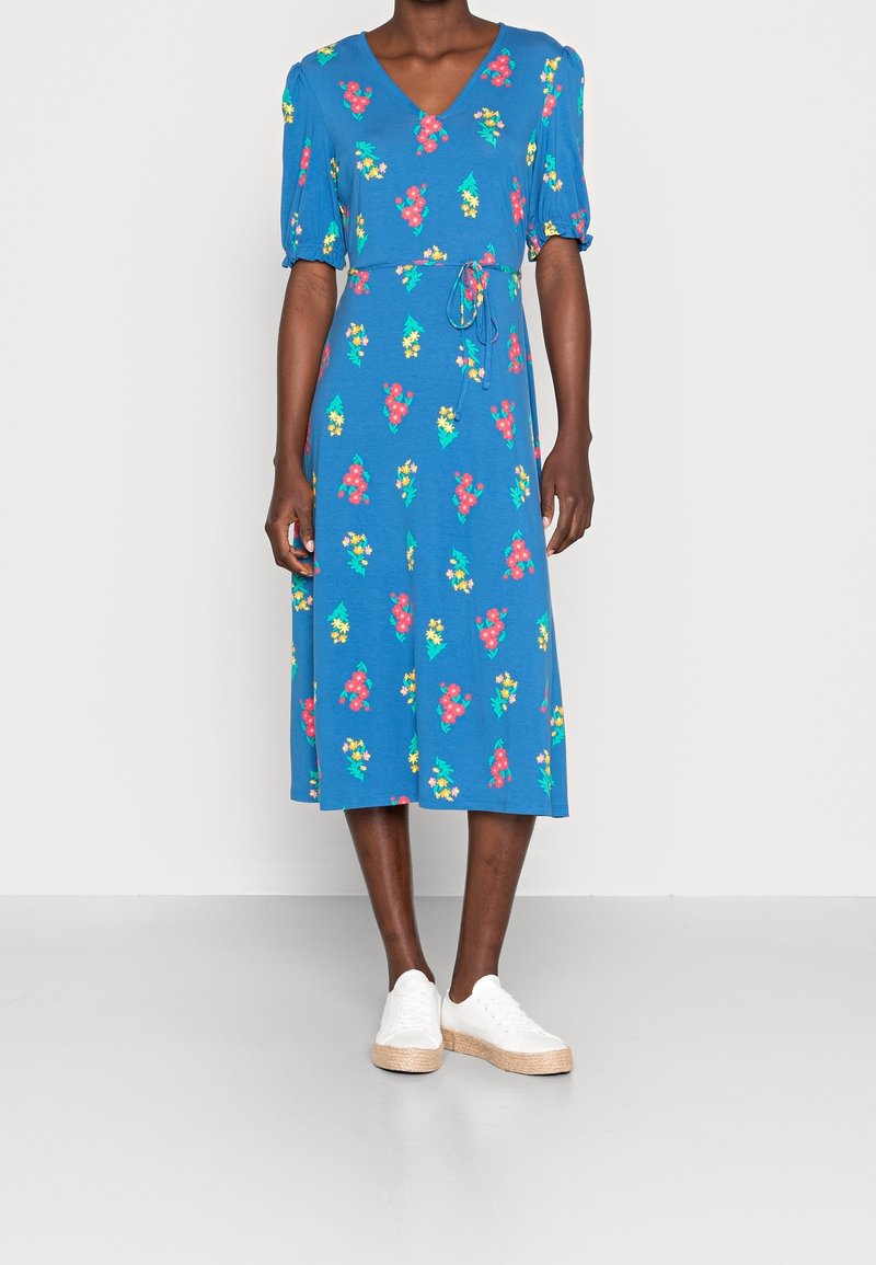 Robe florale bleue avec des manches bouffantes courtes, décolleté en V, taille cintrée et jupe mi-longue. Assortie à des baskets blanches et des semelles compensées en jute.