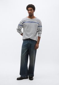 Ριγέ πουκάμισο με μακριά μανίκια σε λευκό χρώμα με μπλε και μαύρες ρίγες, συνδυασμένο με φαρδιά ξεβαμμένα μπλε τζιν και μαύρα παπούτσια. Ορατές είναι οι απλές λεπτομέρειες σχεδίασης.