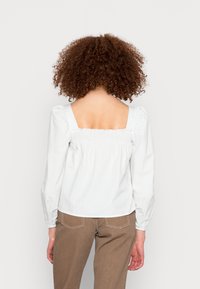 Blusa branca com riscas, mangas bufantes, decote quadrado e detalhe franzido nas costas, combinada com calças castanhas com bolsos.