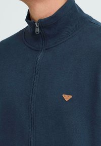 Tumši zilas krāsas jaku ar rāvējslēdzēja aizdari un augstu apkakli, ar mazu brūnu trīsstūra logotipu uz krūtīm un ribota detaļām ap apkakli.