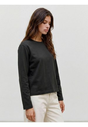 Jeune femme aux longs cheveux ondulés portant un t-shirt noir à manches longues et un pantalon clair, debout devant un fond clair uni.