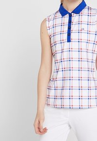 Ärmlös polo tröja i vitt med en blå krage. Har ett rutigt mönster i röd och blå. Tillverkad av ett lent, lätt tyg.