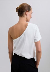 Weißes Baumwoll-T-Shirt mit asymmetrischem Design, einem freiliegenden Schulter und kurzen Ärmeln, kombiniert mit schwarzen Hosen.