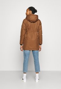 Parka de color marrón, aislada, con capucha, cintura ajustada y mangas largas. Combinada con vaqueros cortos de color azul claro y zapatillas blancas.