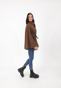 Brązowy poncz z wełny z wysokim kołnierzem i czarnym paskiem, sparowany z niebieskimi skinny jeansami i czarnymi botkami na platformie.