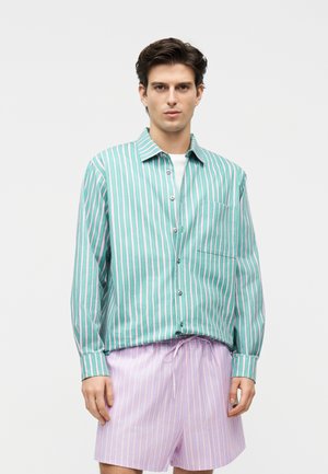 Joven con camisa de botones a rayas en tonos turquesa y blanco sobre una camiseta blanca, y pantalones cortos a rayas morado claro, posando contra un fondo liso.