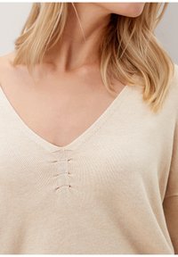 Beige Strickpullover mit einem V-Ausschnitt und drei horizontalen Nahtakzenten auf der Brust. Weicher Stoff, lockere Passform, keine Muster.