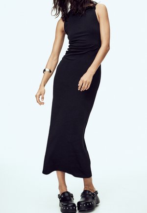 Femme portant une robe midi noire sans manches, des sabots noirs cloutés et un bracelet en or sur le bras gauche sur un fond uni.