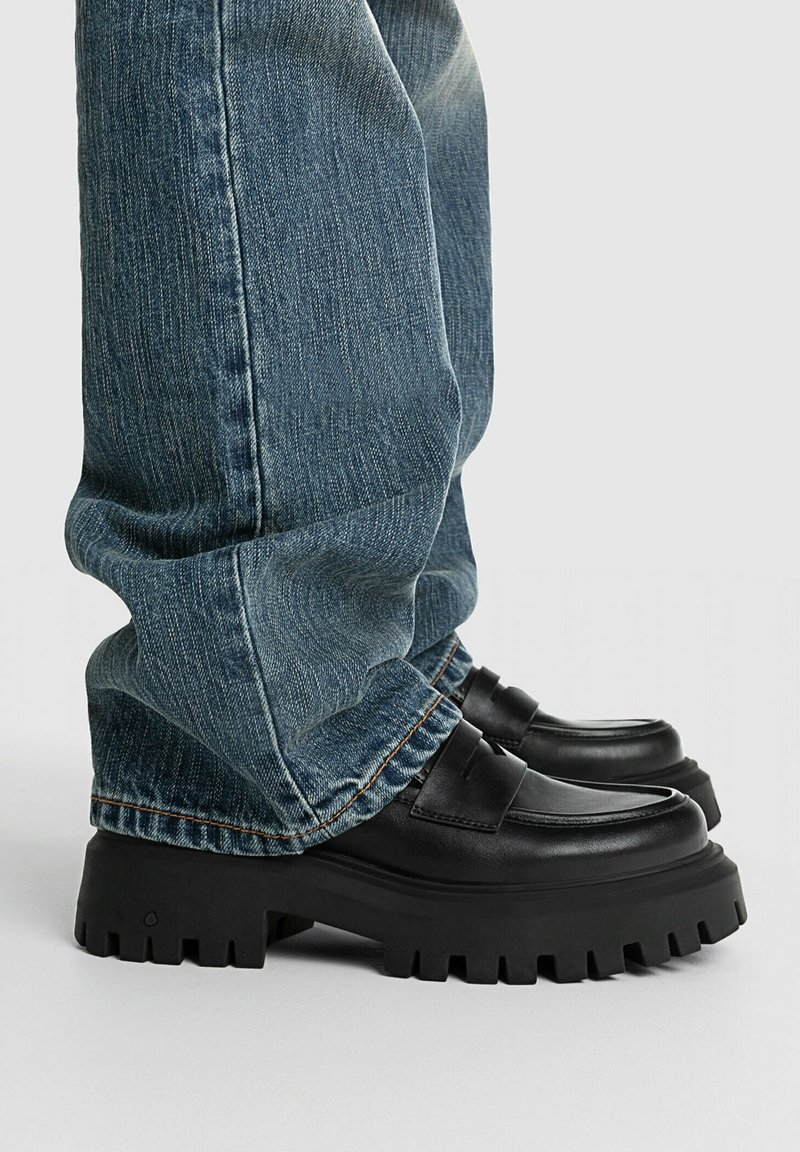 Personne portant un jean en denim bleu et des mocassins en cuir noir épais avec des semelles épaisses à crampons, debout sur une surface claire.
