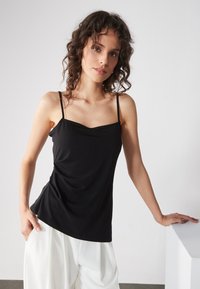 Top nero senza maniche con scollatura drappeggiata, texture liscia, spalline regolabili e vestibilità comoda, abbinato a pantaloni bianchi larghi.