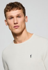 Suéter de cuello redondo en crema claro, hecho de una tela suave, que presenta un pequeño logo en azul marino bordado en el pecho y acabados acanalados en el cuello.