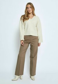 Maglione crema con scollo a V e maniche lunghe, abbinato a pantaloni marroni a vita alta e stivaletti ankle con tacco a blocco beige. Texture liscia visibile.