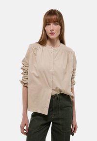 Chemise beige à col rond, avec des détails froncés aux épaules et des manches retroussées, associée à un pantalon vert foncé.
