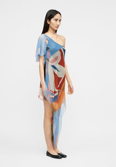 Γυναίκα που φοράει έναν πολύχρωμο, ασύμμετρο φόρεμα με έναν ώμο, από διάφανο ύφασμα και μαύρες επίπεδες γόβες, στέκεται μπροστά σε απλό φόντο.