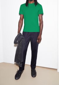 Zelené tričko s krátkým rukávem, tmavé kalhoty, černé strukturované boty a šedá bunda přehozená přes jednu ruku. Jednoduché pozadí.