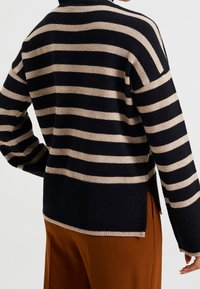 Pruhovaný pletený svetr v námořnické modré a béžové barvě, s volným střihem, ribbovaným lemem a bočními rozparky, vyrobený z měkkého texturovaného materiálu.