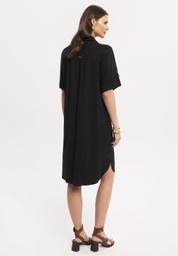 Czarna sukienka koszulowa z krótkimi rękawami, z miękkiego materiału, z bocznymi rozcięciami i zapinaną na guziki kieszenią. Noszona z brązowymi sandałami na słupku.